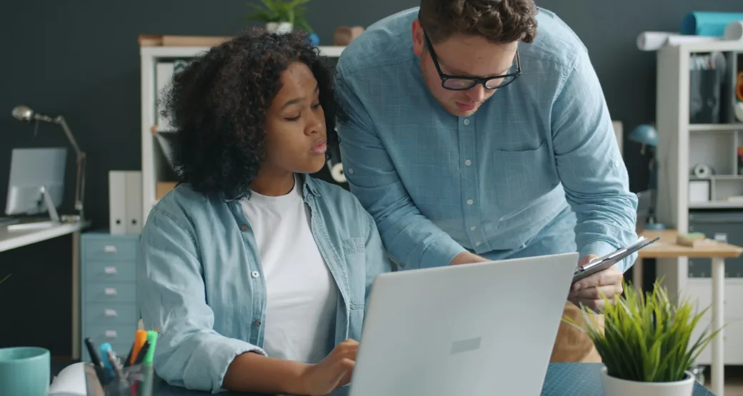 A mentor helps a young professional with her work on a laptop